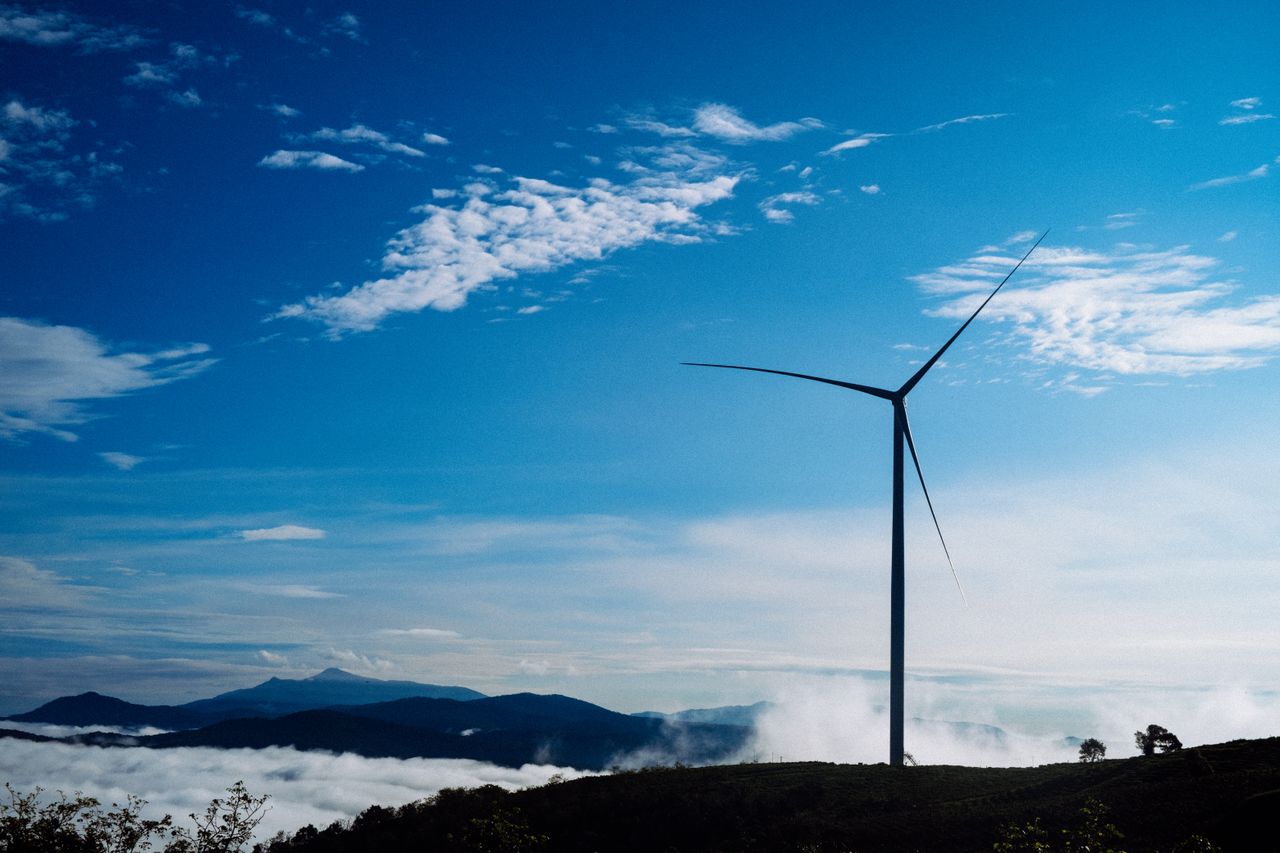 Offerings A solitary wind turbine rises against a vibrant blue sky, surrounded by clouds and distant mountains.