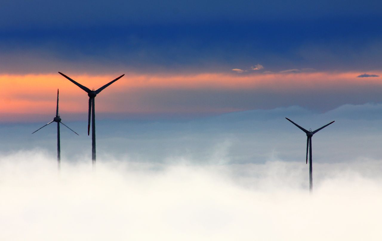 Home Silhouetted wind turbines rise above clouds against a dramatic sunset sky.